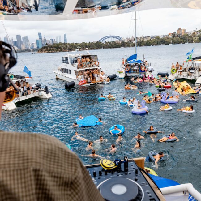 DJ playing music on a boat overlooking a lively scene with people swimming and lounging on inflatables in the water. Several boats are gathered nearby, and a city skyline with a bridge is visible in the background.