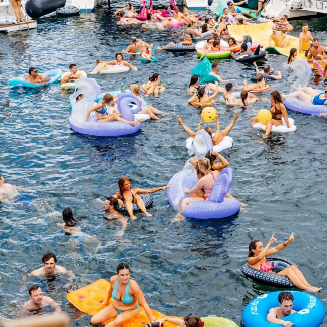 A lively scene of people enjoying a sunny day in a lake or sea, using colorful inflatable floats shaped like swans, unicorns, and rings. Boats are anchored nearby. Participants are relaxing, socializing, and swimming in the clear water.