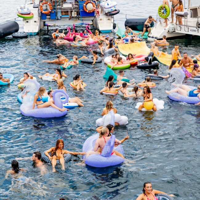 A lively scene of people enjoying a day on a lake, surrounded by inflatable toys and floats in various shapes, such as unicorns and swans. Two boats are docked nearby, and the water is filled with individuals relaxing and socializing.