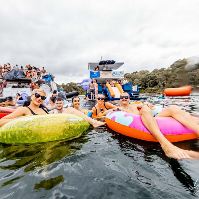 A group of people relax on colorful inflatable rafts in the water near a boat. They are smiling under a cloudy sky, with more people seen on the boat in the background. Trees line the shore, and a design with "Yacht Social" is visible in the corner.