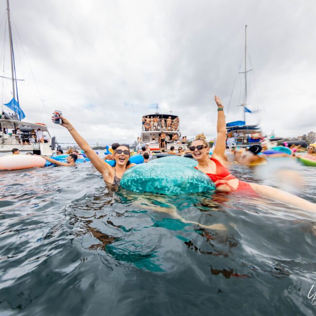 Two people in swimsuits smile and raise their arms while floating on inflatables in the water. Boats are visible in the background, with a cloudy sky above. The scene is lively and social.
