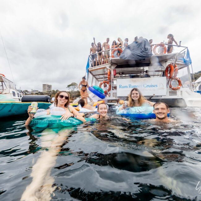 A group of people relax in the ocean, floating on inflatables. They are near a boat crowded with more people. The scene is festive and lively, with several boats in the background. The water is calm, and the sky is cloudy.