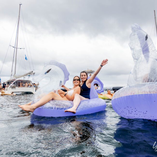 Two people are smiling and relaxing on inflatable unicorn floaties in the water. They are surrounded by several boats and other floaties under a cloudy sky.