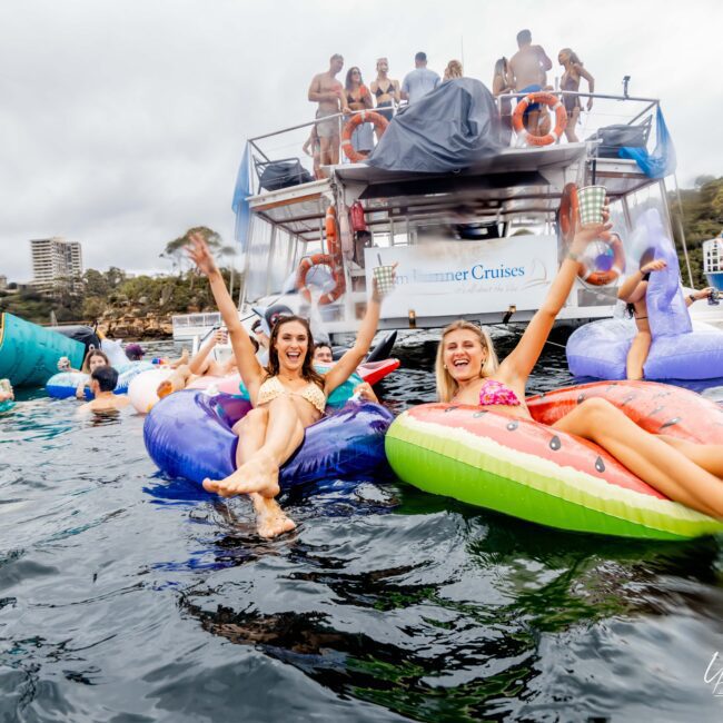 Two people smiling and lounging on inflatable rafts shaped like a star and watermelon slice in the water. Other people are on more inflatables and a boat in the background. The sky is overcast, and the atmosphere is lively and celebratory.