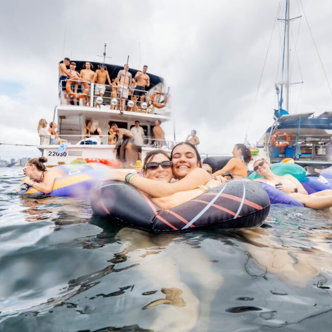 Two people are smiling and floating on an inflatable tube in the water. In the background, a yacht filled with people appears, and another boat is visible to the right. The scene is lively and festive.