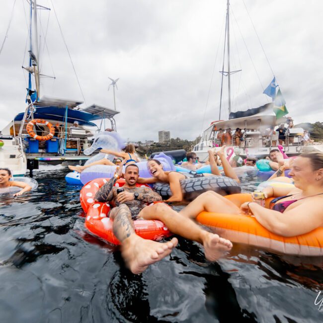 A group of people relax on colorful inflatable floats in the water, enjoying a social gathering near boats. The scene is lively, with laughter and smiles, under a cloudy sky.
