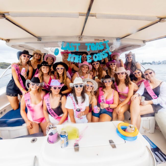 A group of women in pink outfits and party accessories are gathered on a boat, smiling and posing for a photo. They're celebrating with a banner that reads "Last Fling Before the Ring." The background shows a waterway and a distant city skyline.