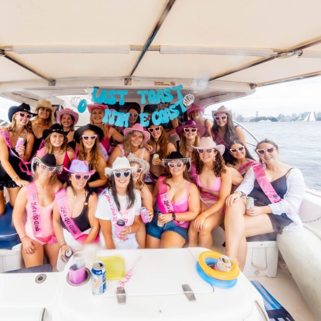 A group of smiling people on a boat celebrating a bachelorette party. They are wearing pink sashes and cowboy hats. A sign above reads "Last Toast Before the Coast." They are docked with scenic water and city skyline in the background.