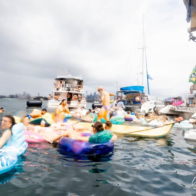 People are having fun on inflatable floats in the water near several yachts. The sky is cloudy, and a cityscape is visible in the background. The atmosphere is lively with multiple boats and people enjoying a day out on the water.