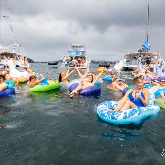 A group of people relax on colorful floaties in the water, surrounded by boats. Some individuals hold hands, while others lounge and chat. The sky is cloudy, and the water is calm.