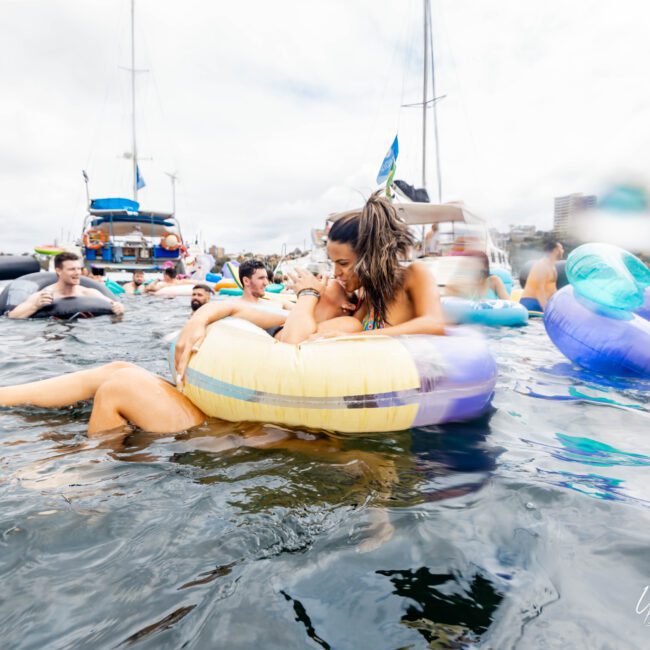 People enjoying a party in the water, with a woman laughing and lying on a yellow inflatable pool float. Others float and swim nearby under a cloudy sky, with boats in the background.
