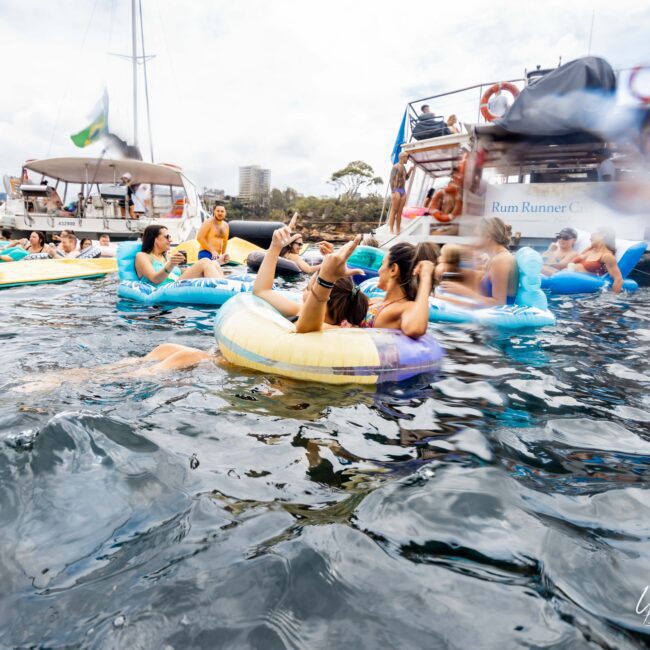 People enjoying a day on the water, floating on inflatable rings and swimming near two anchored yachts. The atmosphere is festive, with socializing and relaxed interactions. Trees and buildings are visible in the background.