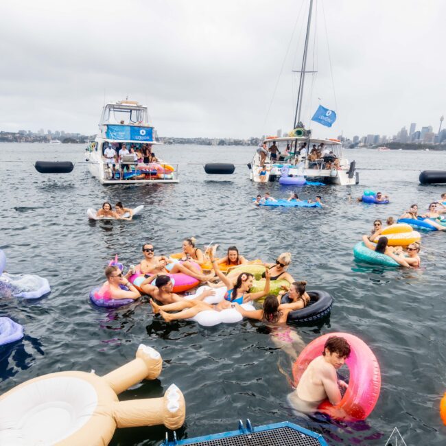 A group of people relaxing on colorful inflatables in a body of water, surrounded by anchored boats. The sky is overcast, and city skyline is visible in the background. Some people are socializing while others float nearby.