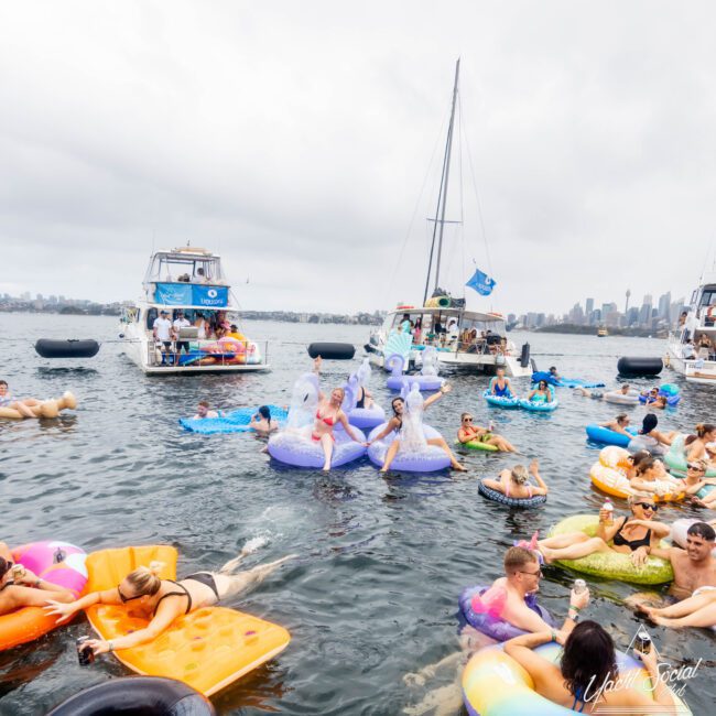 A lively scene on the water with people enjoying a float party. Various colorful inflatables surround several boats. Attendees in swimwear relax and socialize amid a backdrop of a cloudy sky and distant city skyline.