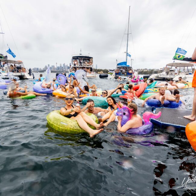 A group of people enjoying a party on inflatable rafts in the water, surrounded by boats. They are wearing swimwear and appear to be having fun, with a cloudy sky and a city skyline in the background.