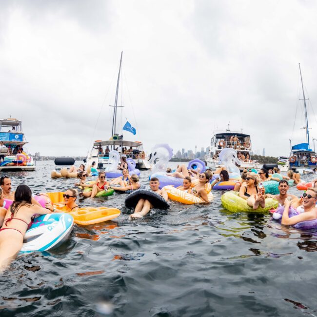 A large group of people relaxing on colorful inflatable floats in the water near several anchored boats. The cloudy sky and distant city skyline are visible in the background. The scene is lively and festive.