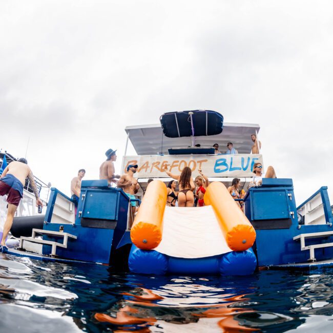 A group of people enjoying a day on a boat named "Barefoot Blue." The scene features inflatable slides extending into the water, and several individuals are swimming or relaxing nearby. The sky is overcast, and trees are visible in the background.