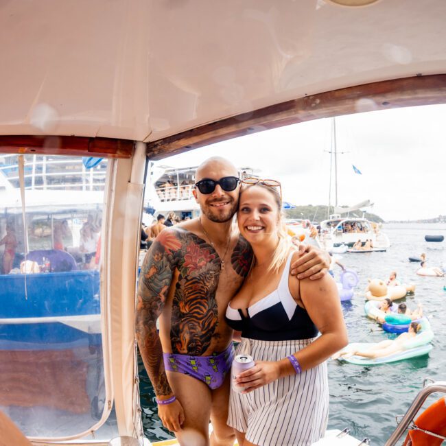 A man with tattoos, wearing purple swim trunks, stands next to a woman in a black and white swimsuit holding a drink. They are smiling on a boat, with a lively group of people and more boats visible on the water in the background.