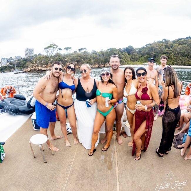 A group of people in swimwear posing on a boat. They are smiling and holding drinks. Trees and buildings are visible in the background. The logo "Yacht Social Club" is on the bottom right corner of the image.