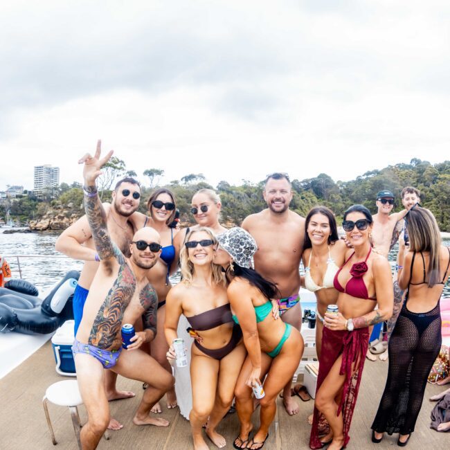 A group of people in swimwear pose and smile on a boat. There are drinks in hand, with a cloudy sky and trees in the background. One person is making a peace sign.