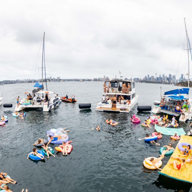 A party on the water with people on colorful inflatables surrounded by several boats. The boats have groups of people and the city skyline is visible in the background. The sky is overcast.