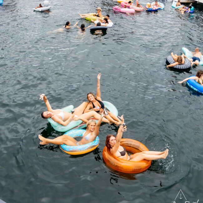 A group of four people joyfully float on colorful inflatable tubes in a lake, raising their arms playfully. Other people are seen on various inflatables in the background, and boats are nearby.