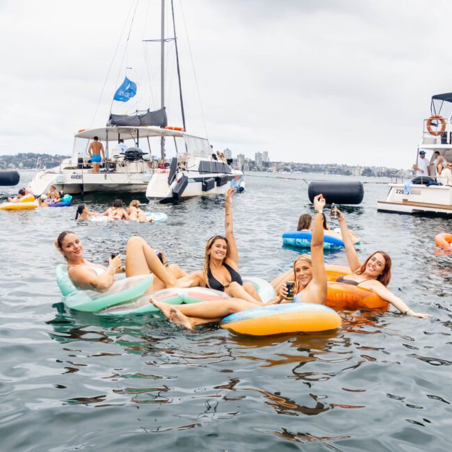 Four people are relaxing on colorful inflatable loungers in the water, holding up drinks and smiling. In the background, several boats and more people enjoying the water are visible under a cloudy sky.