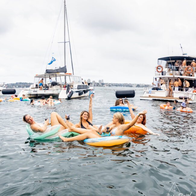 Four women are relaxing on colorful inflatable rings in the water, holding drinks and smiling. Behind them are sailboats and yachts with other people enjoying the day. The sky is cloudy, and the scene is lively and festive.