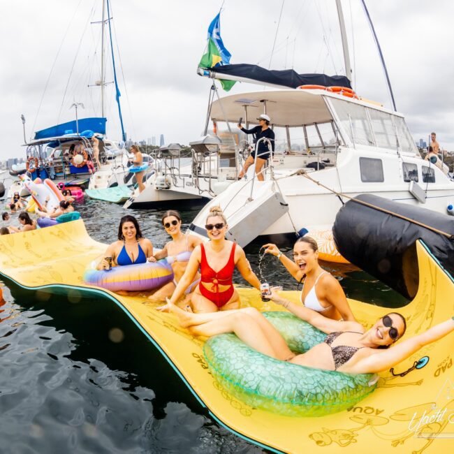 Five people are laughing and lounging on inflatable floats in the water near two yachts. The group enjoys a festive atmosphere, with more people and colorful floats in the background. The sky is overcast.