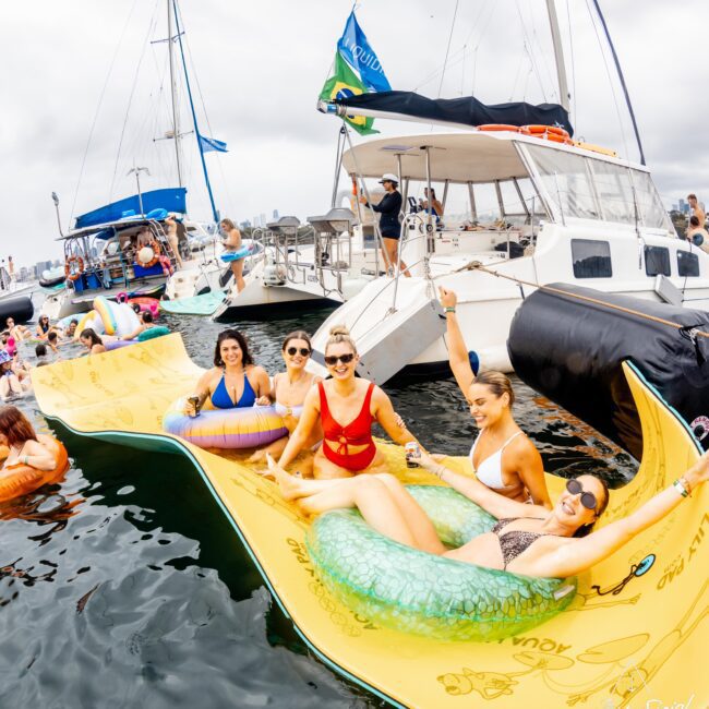 A group of people relaxes on various floats in a marina, enjoying a sunny day. Several boats are docked nearby. Some are holding drinks and wearing swimsuits. The atmosphere is lively and social. Flags can be seen in the background.