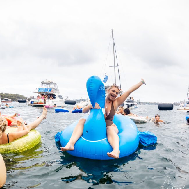 A person joyfully riding a large blue inflatable toy on water, surrounded by others on various inflatables. Boats are in the background under an overcast sky. The mood is festive and lively.