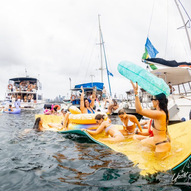 A lively scene of people enjoying a party on the water. Several people are on a floating mat in swimsuits, surrounded by boats. Some are holding inflatable toys, while others relax and socialize. The boats host more people in the background.