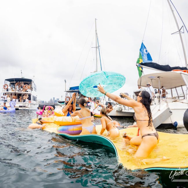 People are enjoying a boat party in sunny weather. They are lounging on inflatable sunbeds and floats in the water, surrounded by yachts. Some are holding drinks and large inflatable toys, creating a lively and festive atmosphere.