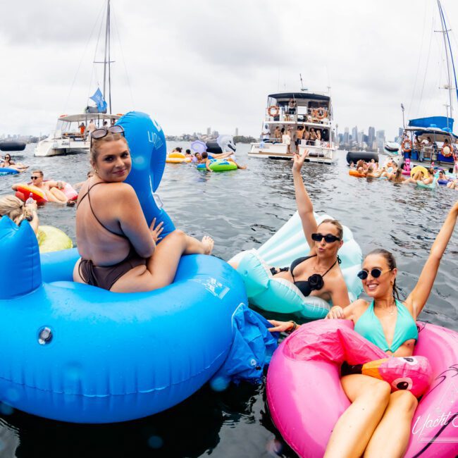People enjoying a pool party on inflatable floats in the water, with boats and a city skyline in the background. The scene is lively with participants smiling and posing under a cloudy sky.
