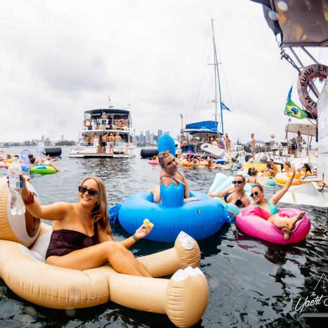People relaxing on inflatable floats in a marina, surrounded by boats. A woman in sunglasses holds a drink, while others lounge nearby. One person wears a Marge Simpson costume. The city skyline is visible in the background.