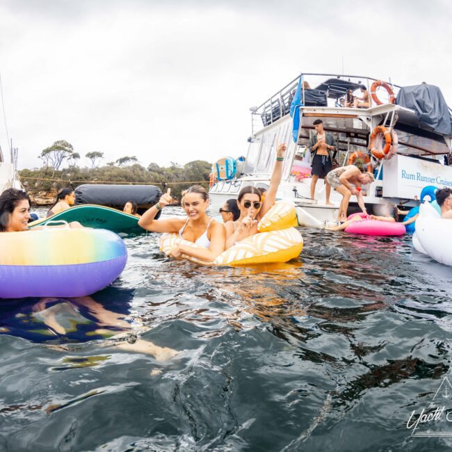 People are enjoying a day on the water, lounging on colorful inflatable floats near boats. The lake or ocean setting is lively with various individuals swimming and relaxing. The atmosphere is casual and fun.