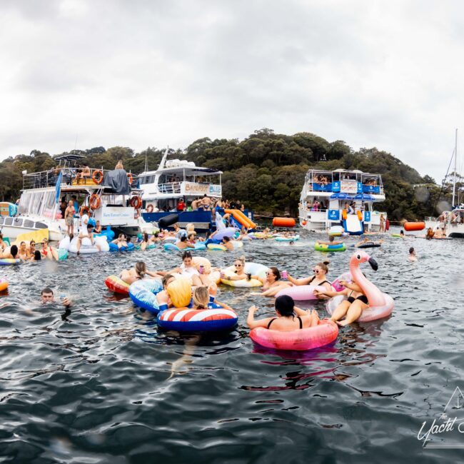 A lively scene on the water with people relaxing on colorful inflatable floats, including a pink flamingo, near several anchored boats. The sky is overcast, and the water is busy with activity and various boats in the background.