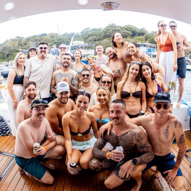 A group of people smiling and posing together on a boat deck. Many are in swimsuits and sunglasses, holding drinks. Trees and other boats are visible in the background. The atmosphere is lively and festive.