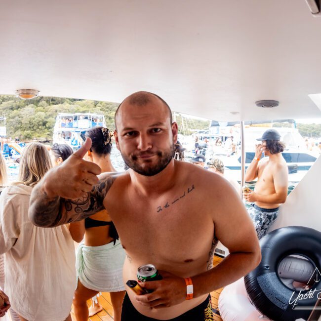 A shirtless man with tattoos gives a thumbs-up while holding a can on a boat deck, surrounded by others in swimwear. There are inflatables nearby, with several boats and trees in the background.