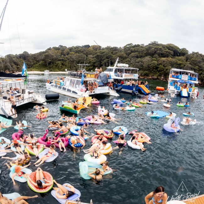 A fun scene of people on colorful inflatable floats in the water near several boats. The setting is a lush, green shoreline, and the group appears to be enjoying a social event under a cloudy sky.