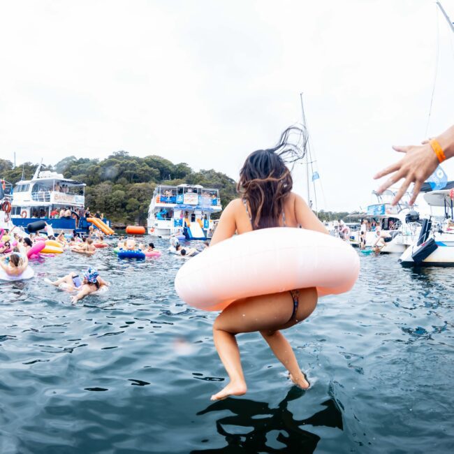 A person wearing a swim ring jumps into a lake, surrounded by other people swimming and floating near several boats. The sky is cloudy, and trees line the background. A person on the right gestures as if encouraging the jump.