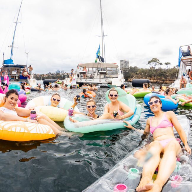 A group of people relax on colorful inflatable floaties in the water, holding drinks. Sailboats are anchored nearby under a cloudy sky. Everyone appears to be enjoying the social event.