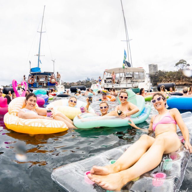A group of people relaxing on colorful pool floats in a river or lake. They're holding drinks and smiling at the camera. Boats are anchored in the background under a cloudy sky. The atmosphere is lively and festive.