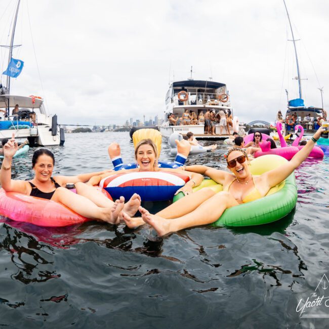 Three people enjoying a day on the water, lounging on colorful inflatable pool floats. They are surrounded by boats and other people in the background, all having fun under a cloudy sky.