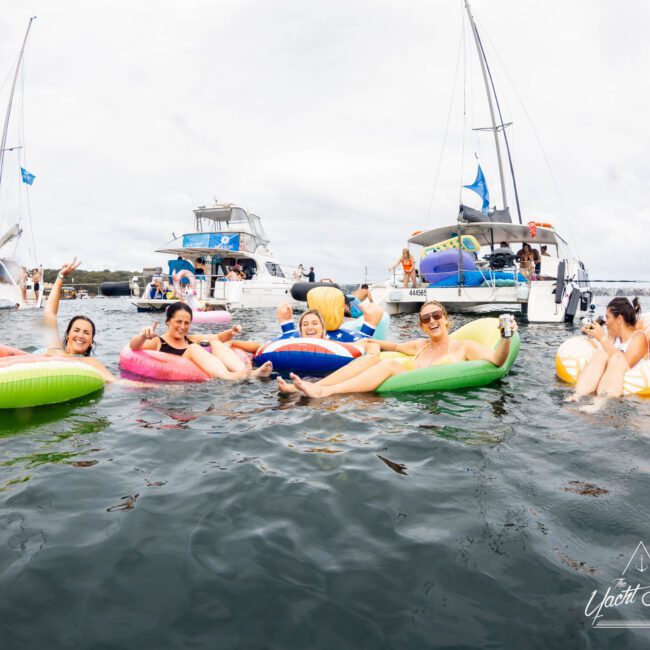 A group of people lounging on colorful inflatable pool floats in the water, with several boats and yachts visible in the background. They appear relaxed and are smiling, enjoying a day on the water. The sky is overcast.