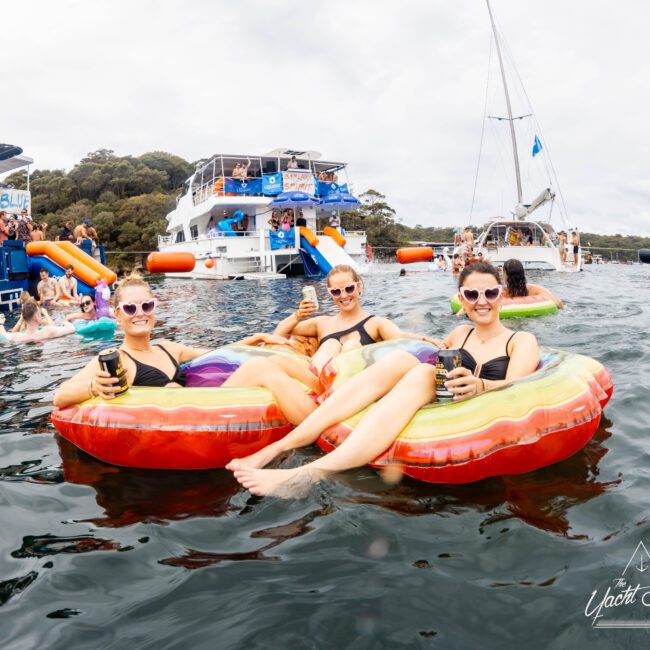 Three people in swimsuits relax on inflatable floaties in the water, holding drinks and smiling. Behind them, a boat with people on board and a sailboat are visible. It's a cloudy day.