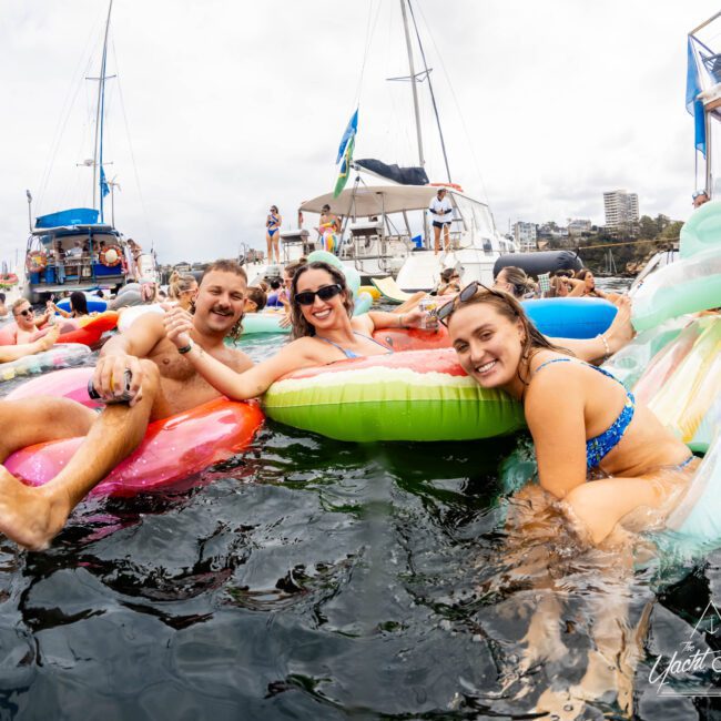 A group of people in swimsuits relax on colorful inflatables in the water, surrounded by yachts. They are smiling and enjoying a sunny day. In the background, more people and boats are visible. The setting is lively and festive.