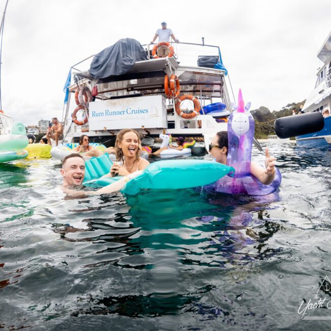 People enjoying a boat party in the water, surrounded by colorful inflatables, including a purple unicorn. Smiles and laughter abound as they float near the boat "Rum Runner Cruises." Several boats are seen in the background.