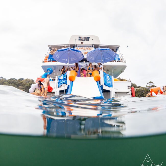 People enjoying a boat party, seen from a water-level perspective. The boat has slides, and several individuals are using them. Blue umbrellas provide shade on the deck, and the Yacht Social Club logo is visible in the photo. Trees are in the background.