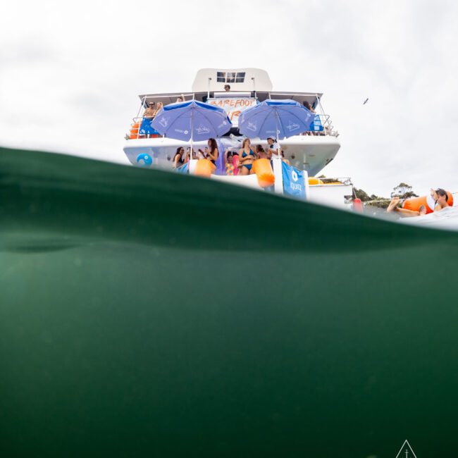 Partial view of a yacht with people onboard under umbrellas, seen from the waterline. The lower half of the image shows a greenish underwater view. Logo reads "The Yacht Social Club" at the bottom right corner.